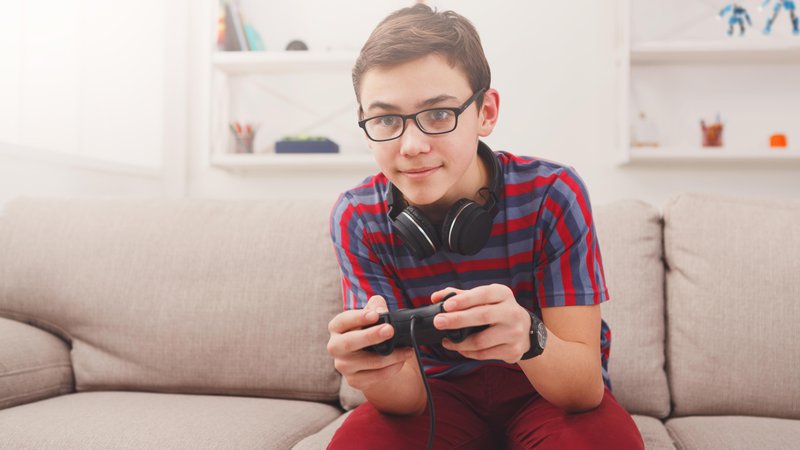 Fotografija: Gaming video games concept - teenage boy playing football game with joystick and headphones, enjoying sitting on sofa in living room at home Foto: Getty Images/iStockphoto
