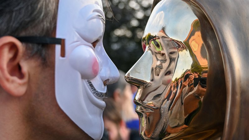 Fotografija: Moški pozira z masko ob bronastem kipu Satoshija Nakamota na Madžarskem. Foto: Attila Kisbenedek / AFP
